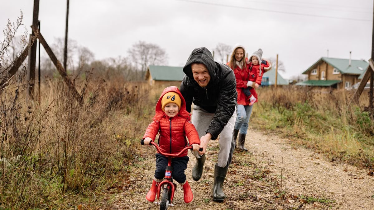 A happy family enjoys a bonding moment outdoors with a child learning to ride a bicycle on a fall day.